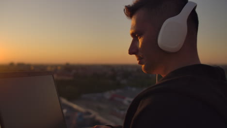 Hipster-man-with-a-laptop-on-the-edge-of-the-roof.-Freelancer-at-work.-Wireless-mobile-Internet.-He-works-on-the-Internet-with-headphones-and-listening-music